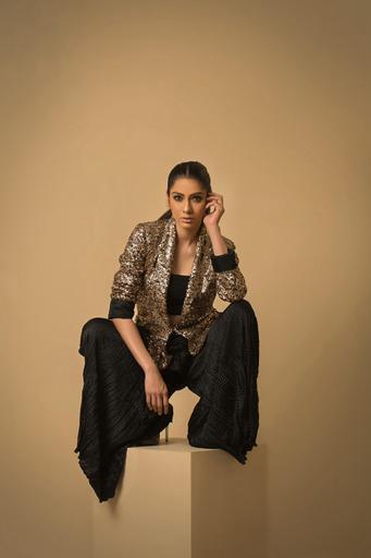 Woman in a gold sequin jacket and black pants sitting on a beige pedestal against a beige background