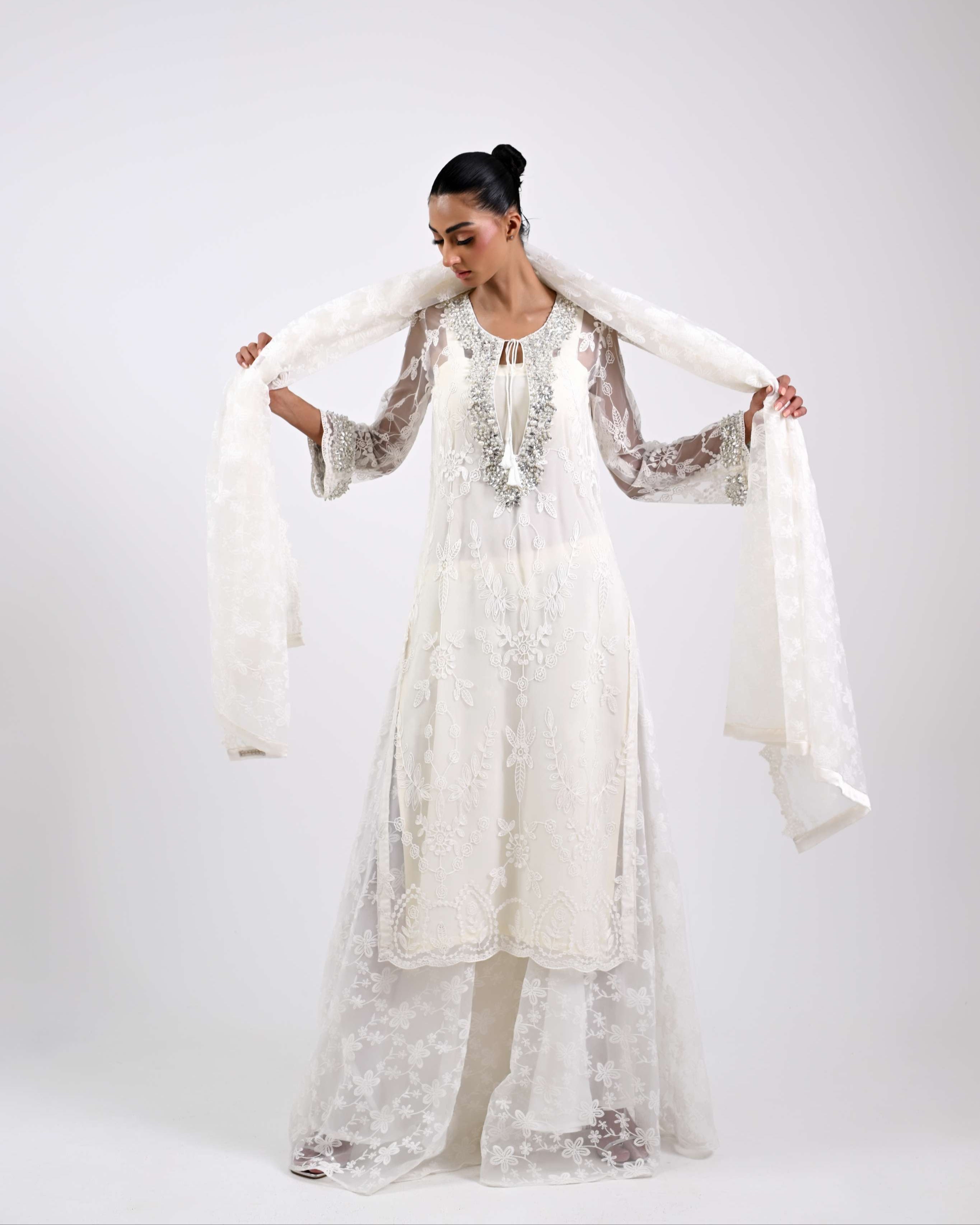 Woman wearing a long, light-colored embroidered dress with a matching shawl against a white background