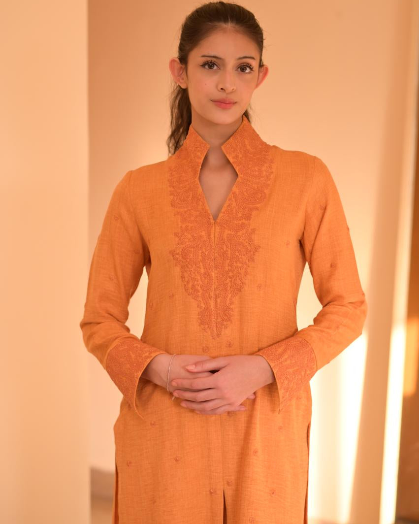 Woman wearing an orange traditional outfit with intricate designs against a warm-toned background